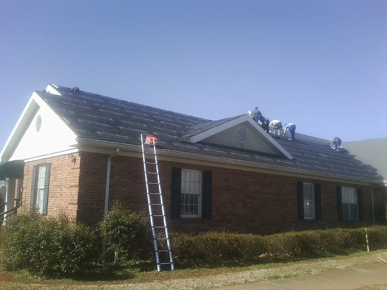 Crew tearing shingles off a brick ranch roof with a ladder set — Emergency Roof Repair work by our crew in Kitty Hawk